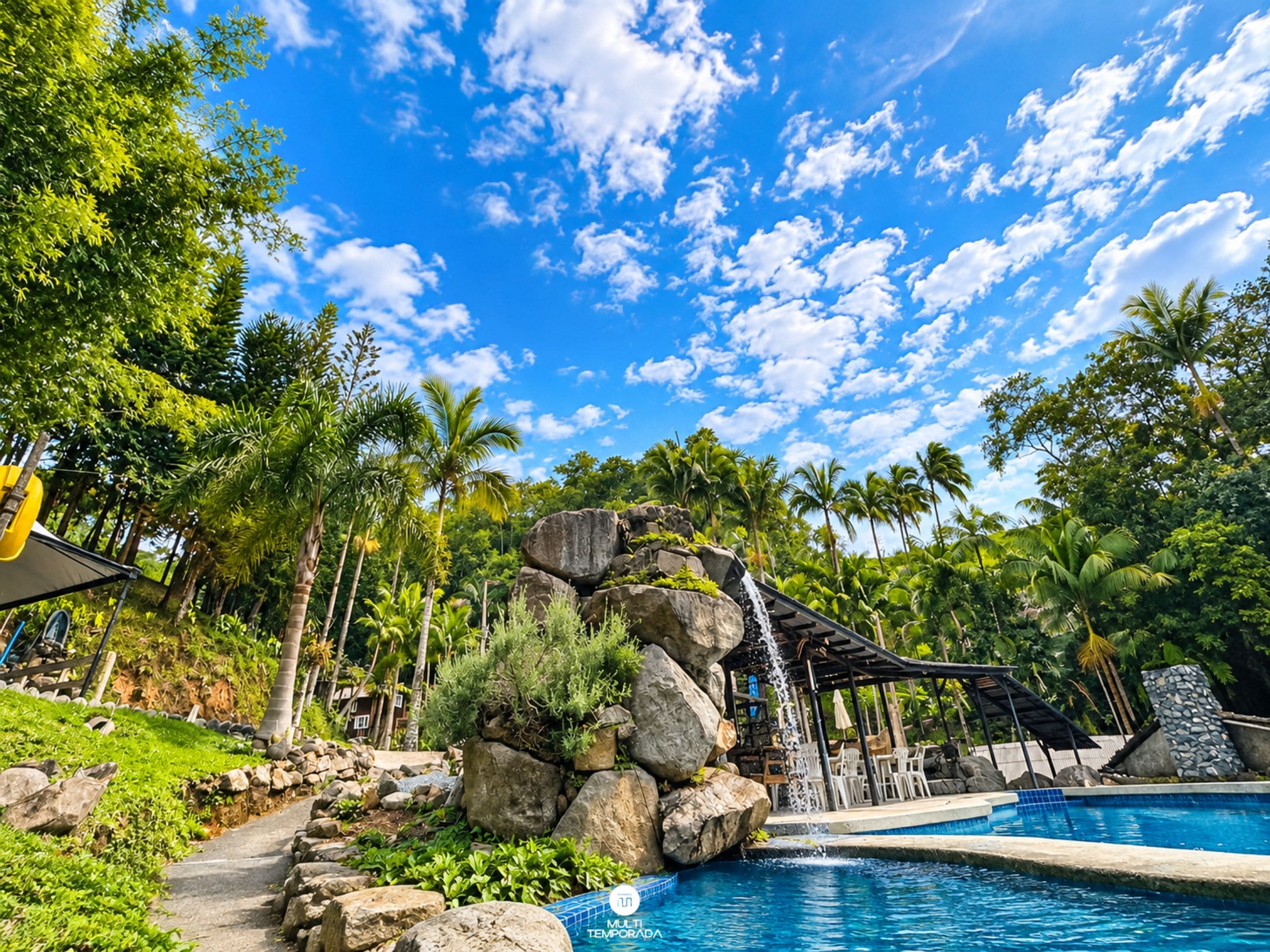 Área de piscina com cachoeira, cercada por vegetação tropical e céu azul límpido.