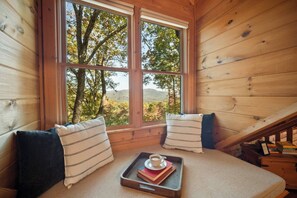 Habitació d'una cabana acollidora amb un finestra gran, una cadira amb coixins i un safata amb llibres i una tassa.