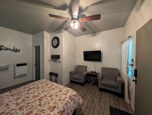Een slaapkamer met een bed, twee fauteuils, een plafondventilator, een aan de muur bevestigde televisie, een klok en een raam met zonwering.