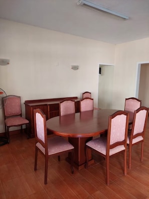 Ein Esszimmer mit einem runden Tisch, sechs Stühlen, einer kleinen Kommode und einem Standventilator.