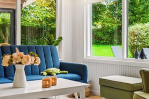 A room with a blue sofa, a white table, and a view of a green garden through large windows. - Lovely holiday home in Dalby Huse (Jægerspris)