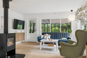 A modern living room with a fireplace, a blue sofa, a white coffee table, and a large window with a view of greenery. - Lovely holiday home in Dalby Huse (Jægerspris)