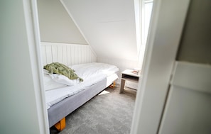 Une petite pièce blanche, avec un toit en pente, comprenant un lit simple, une table de chevet et une fenêtre.