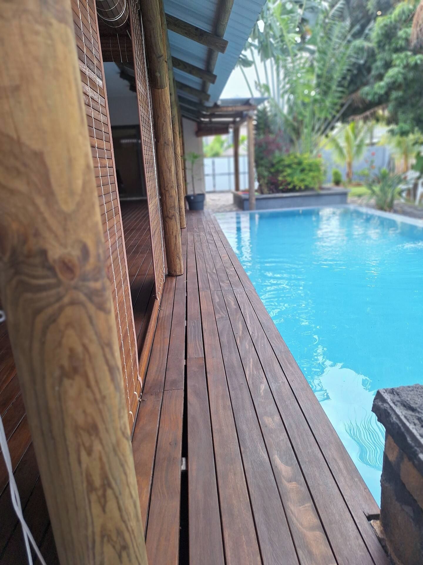 A wooden deck leading to a swimming pool with a covered area and greenery in the background.
