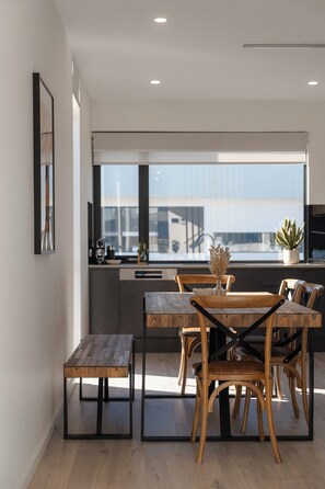 A modern kitchen with a dining area, featuring a wooden table and chairs, a small bench, and a view of the cityscape through large windows. - Maison Central | 3BR 3 Bath + Parking | CBD Luxe (Adelaide)