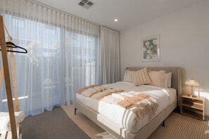 A bedroom with a large bed, a bedside table with a lamp, sheer curtains, and a framed picture on the wall. - Maison Central | 3BR 3 Bath + Parking | CBD Luxe (Adelaide)