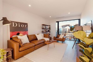 A modern living room with a brown leather sofa, a coffee table, and a bookshelf. There is a large poster on the wall and a view of the outside through a large window. - Maison Central | 3BR 3 Bath + Parking | CBD Luxe (Adelaide)