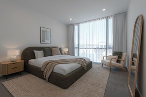 Ein modernes Hotelzimmer mit einem großen Bett, einem Nachttisch mit Lampe, einem Sessel und Blick auf die Stadt durch große Fenster.
