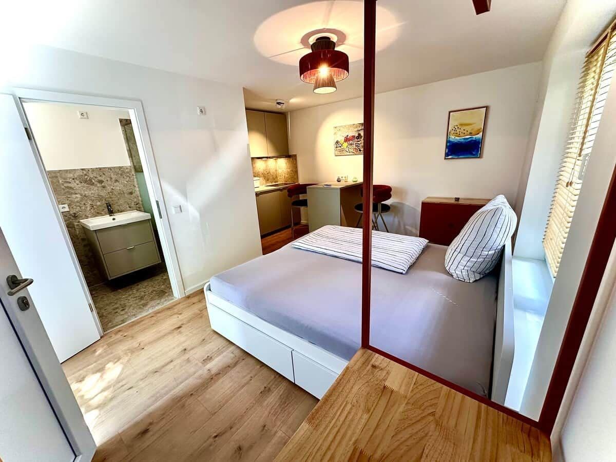 A bedroom with a bed, a bedside table, a mirror, a window with blinds, and a door leading to a bathroom.