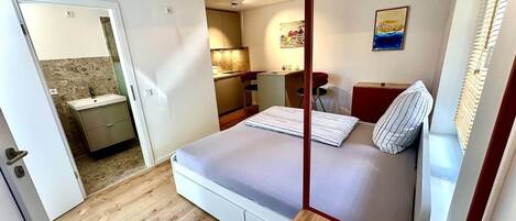 A bedroom with a bed, a bedside table, a mirror, a window with blinds, and a door leading to a bathroom.