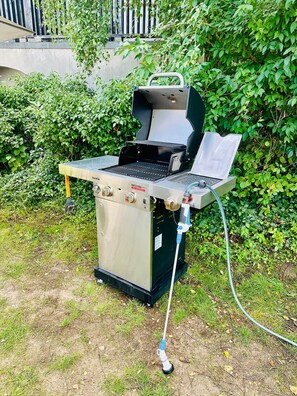 An outdoor grill with a control panel and a hose, set against a backdrop of greenery and a concrete structure. - Stylish Lakefront Studio in Caputh with Lake Access & Garden (Schwielowsee)