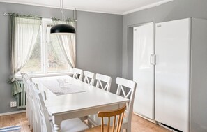 Ein Essbereich mit einem weißen Tisch und Stühlen, einer schwarzen Pendelleuchte, einem Fenster mit Vorhängen und einem weißen Kühlschrank.