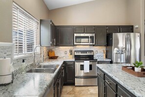 A modern kitchen with dark cabinets, stainless steel appliances, a marble countertop, and a window with blinds. - 3BR Phoenix Retreat Near Golf & Water Parks (Phoenix)