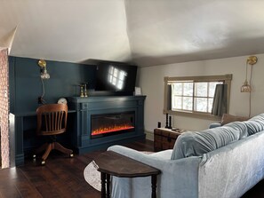 A cozy living room with a fireplace, a television mounted above it, a comfortable sofa, and a window with curtains. - Enjoy a historical carriage house, newly renovated with modern amenities! (Plainwell)