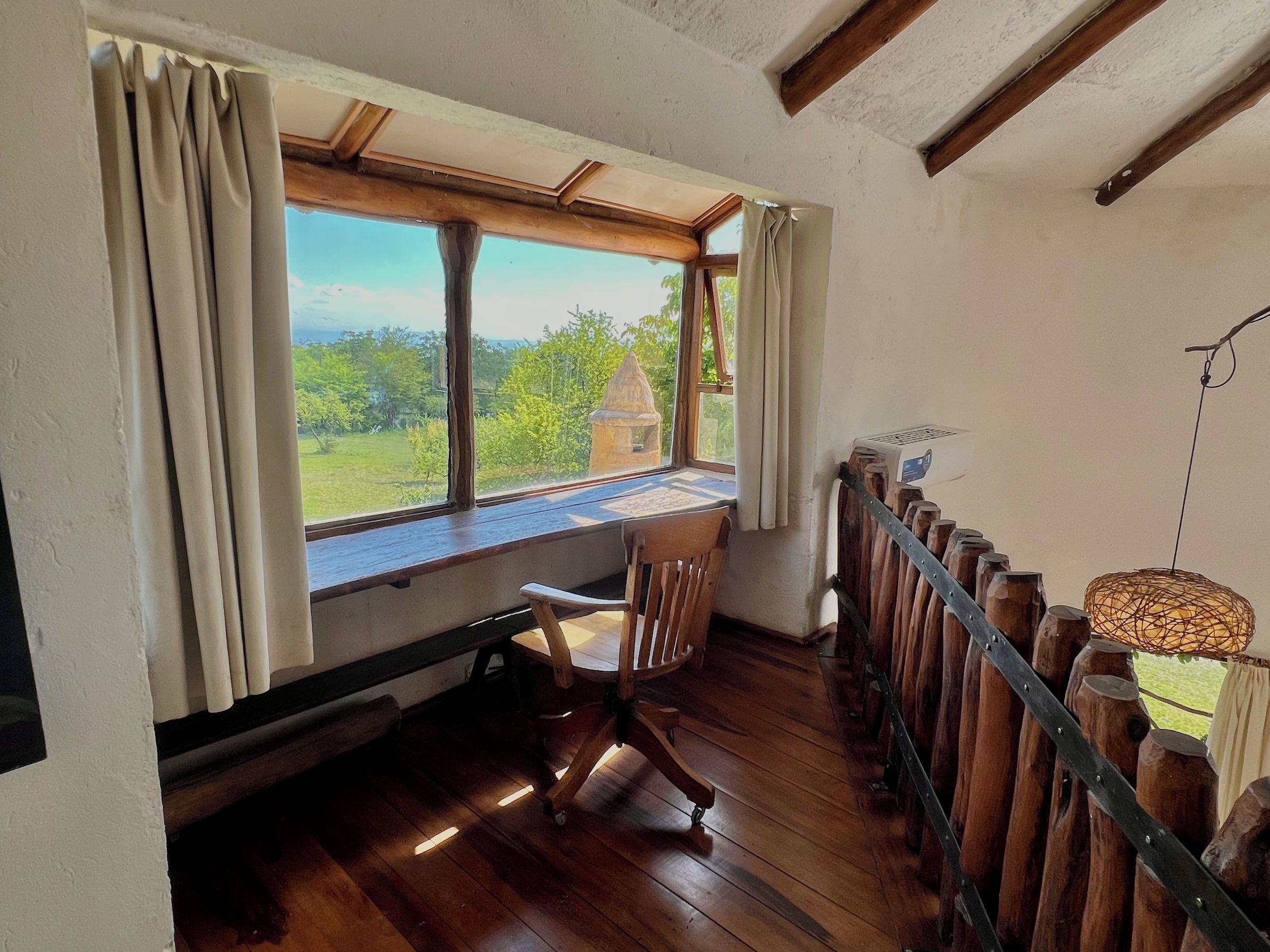 A room with a wooden desk, a chair, a view of greenery and a building outside, and a hanging light fixture.