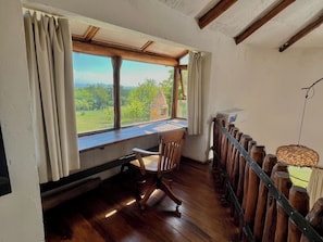 A room with a wooden desk, a chair, a view of greenery and a building outside, and a hanging light fixture.