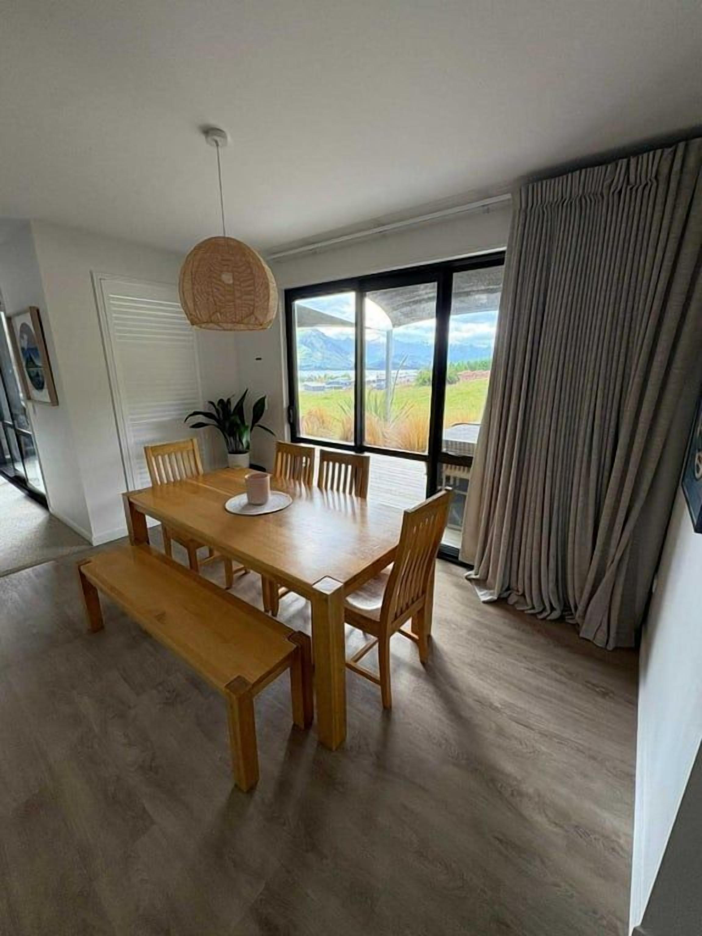 A dining area with a wooden table and benches, large windows with curtains, and a view of the outdoors.