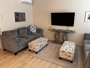 A living room with a grey sofa, two patterned ottomans, a flat-screen TV, and a small table with a remote and a book. - NEW Enjoyable 2-bedroom townhome in Twin Falls (Twin Falls)