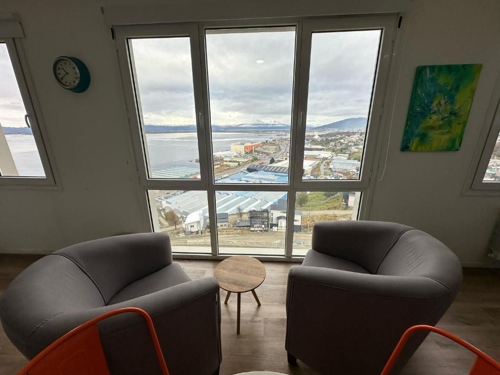 A room with large windows, two grey armchairs, a small wooden table, and a clock on the wall.