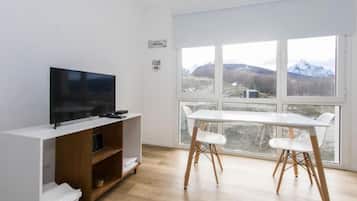 Una habitación moderna con una televisión de pantalla plana sobre un soporte de madera, una mesa blanca con dos sillas y un ventanal con vistas a la montaña.
