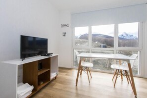Een moderne kamer met een flatscreen televisie op een houten standaard, een witte tafel met twee stoelen en een groot raam met uitzicht op de bergen.