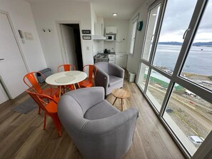 Een moderne kamer met een eethoek, een grijze fauteuil en uitzicht op zee door grote ramen.