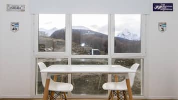 Habitación con un ventanal amplio que ofrece vistas a montañas y un edificio distante. Hay una mesa blanca con dos sillas al frente.