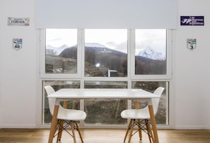 Habitación con un ventanal amplio que ofrece vistas a montañas y un edificio distante. Hay una mesa blanca con dos sillas al frente.