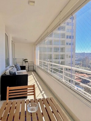 Ein Balkon mit Blick auf eine Stadtlandschaft, einem Holztisch und einer schwarzen Rattan-Couch mit gemusterten Kissen.