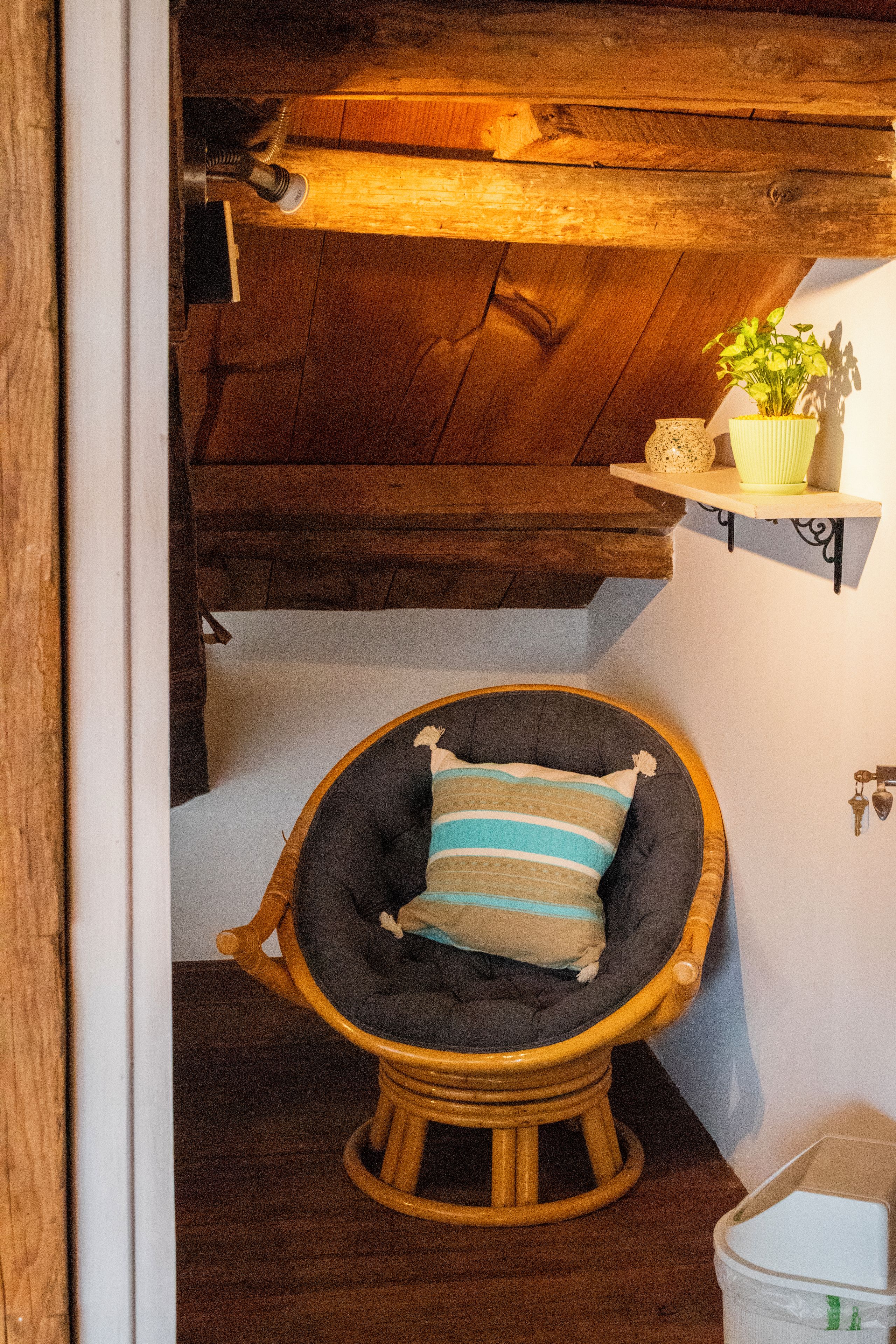 A corner room with a wooden ceiling, a wicker chair with a striped cushion, and a small shelf with a plant.