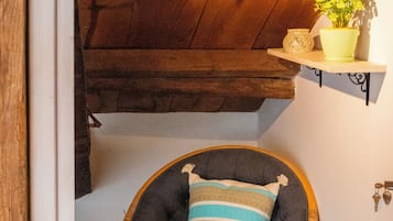 A corner room with a wooden ceiling, a wicker chair with a striped cushion, and a small shelf with a plant.