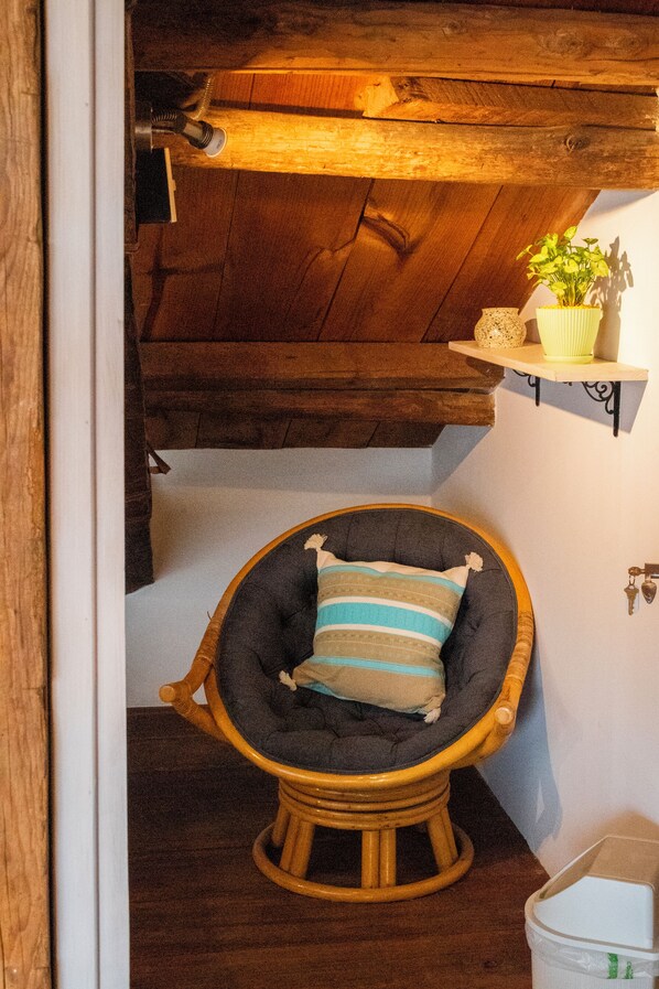 A corner room with a wooden ceiling, a wicker chair with a striped cushion, and a small shelf with a plant.