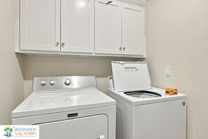 A laundry room with a front-loading washer and dryer, a cabinet above, and a Destination Vacation logo. - 4BR Coastal Retreat | Heated Pool + Pickleball (Gulf Shores)