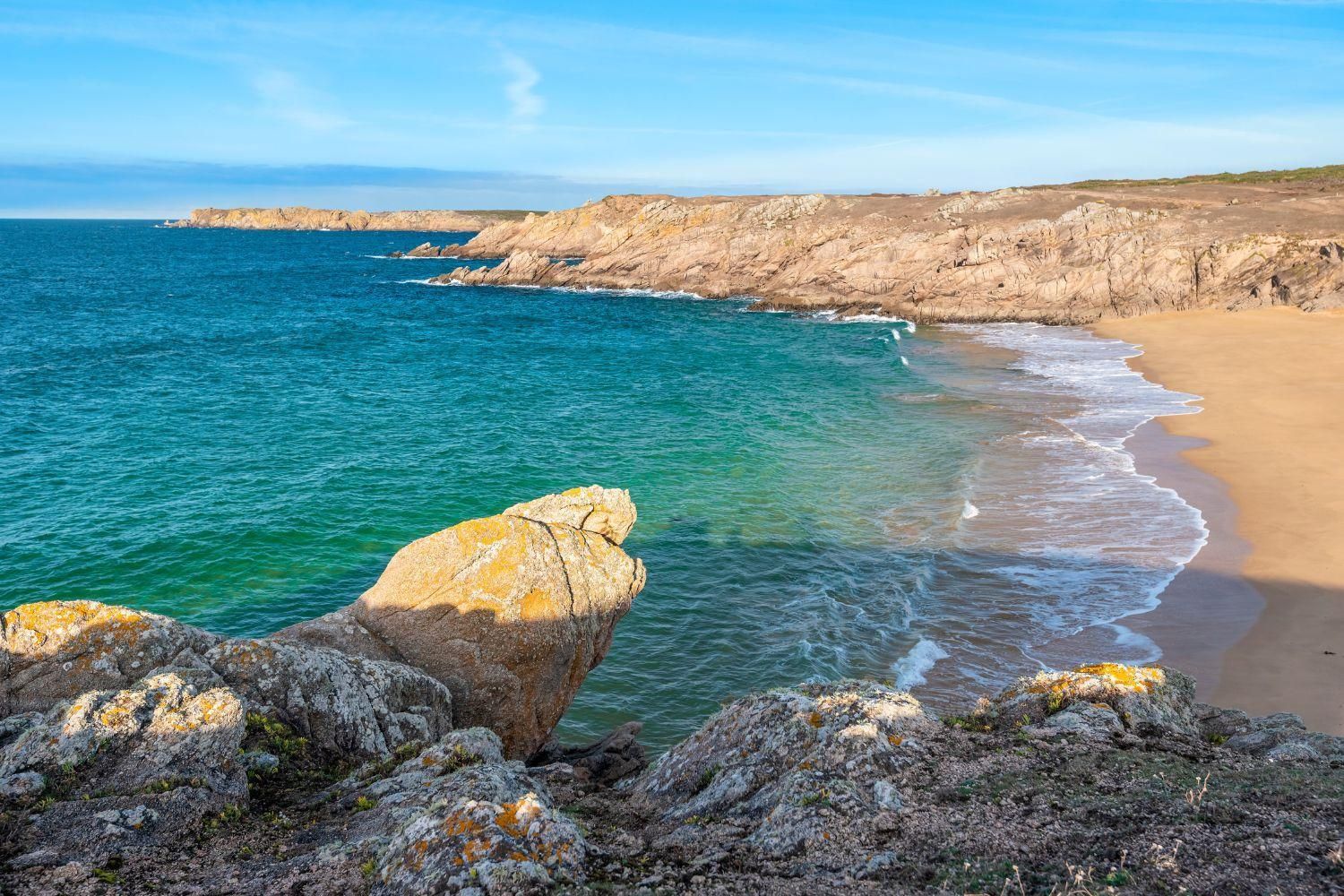 Un paysage côtier caractérisé par des falaises rocheuses, une mer d’un bleu limpide et une plage de sable.