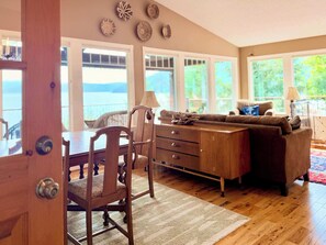 A room with a wooden dining table, chairs, a sofa, and a view of the outdoors through large windows. - Lake Coeur d'Alene 3BR Dock, Kayaks & Boat Slip (Worley)