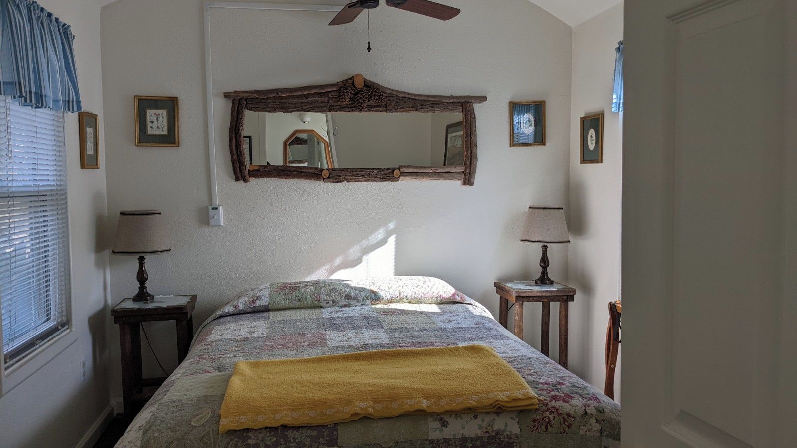 A bedroom with a bed, two bedside tables, a ceiling fan, a large mirror, and framed pictures on the wall.