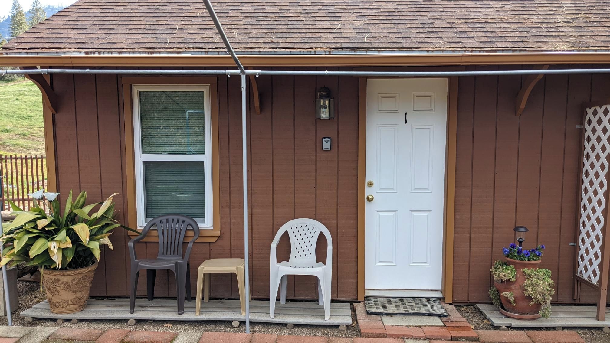 A wooden house with a white door, two plastic chairs, a potted plant, and a metal gate.