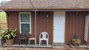A wooden house with a white door, two plastic chairs, a potted plant, and a metal gate. - The Apple Place Room #1 (Miramonte)