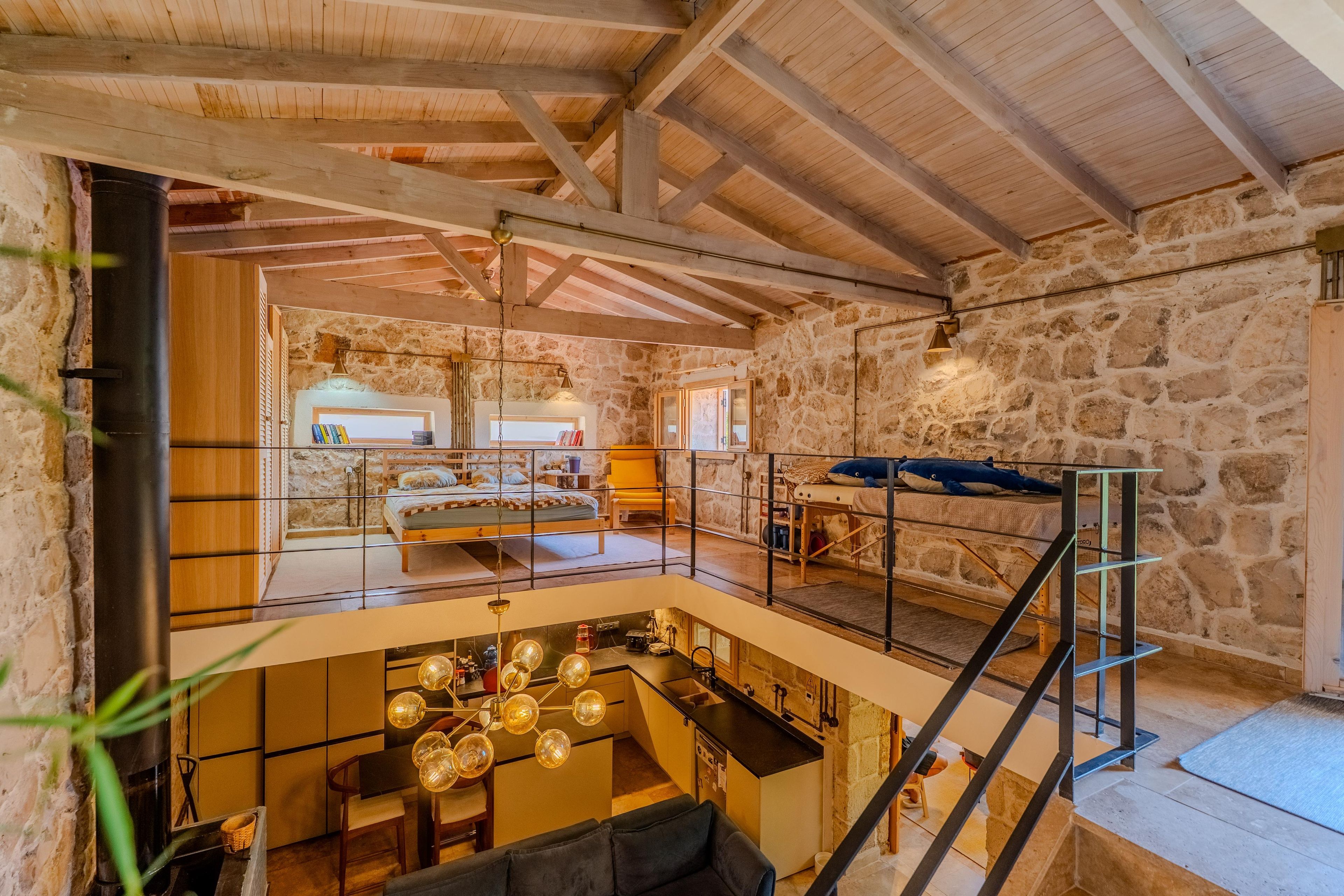 A spacious loft with a high ceiling, exposed wooden beams, a stone wall, a mezzanine level with a bed, and a kitchen area.