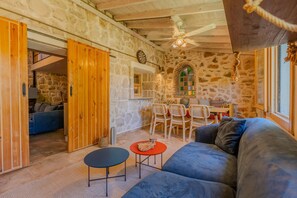 A cozy interior with a stone wall, wooden ceiling, a blue sofa, a small round table, and a dining area with chairs. - The magic of the stone hause,withheated and cooled pool&peaceful escape (İzmir)