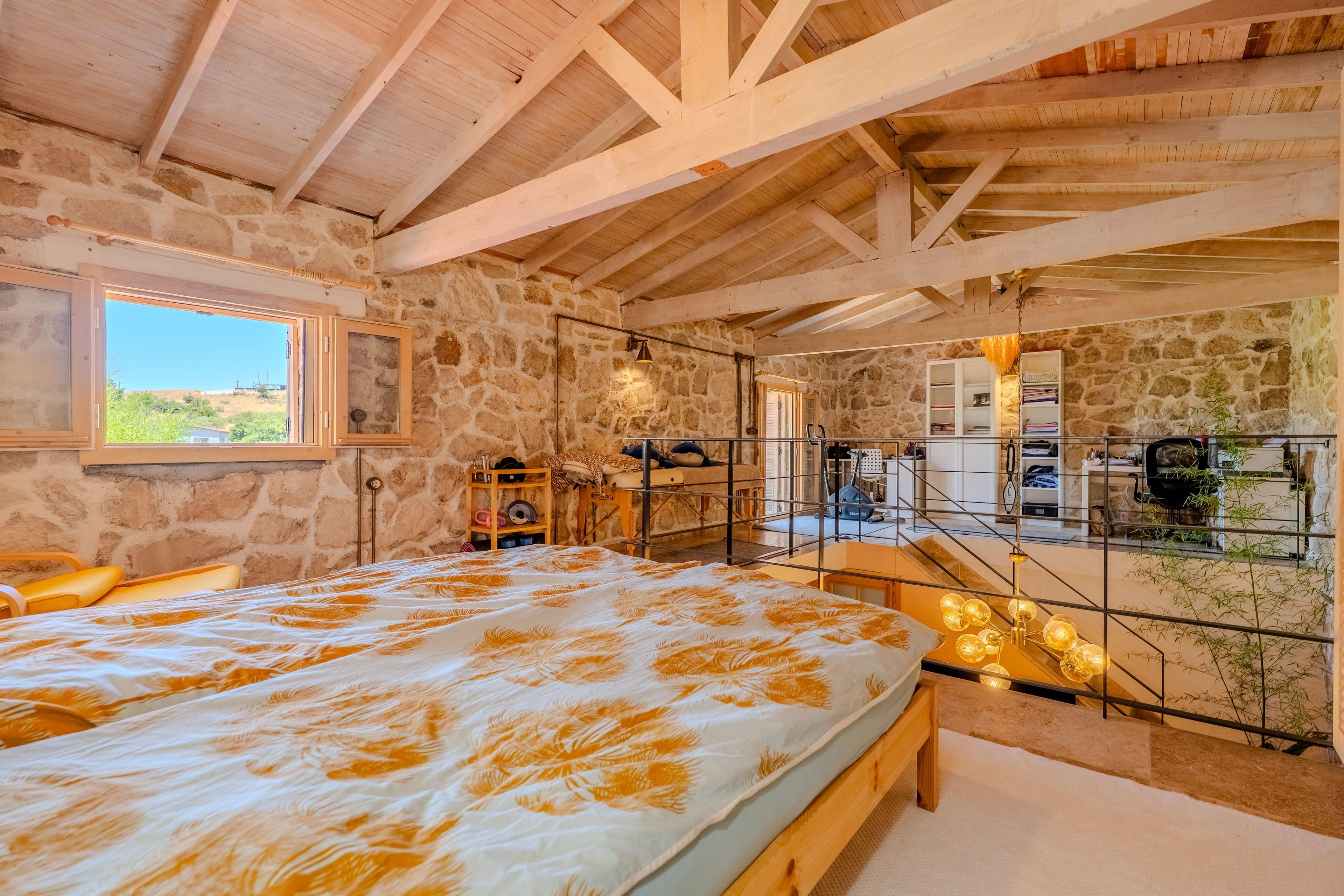 A bedroom with a bed, a window, a stone wall, and a balcony with a view.