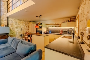 A modern kitchen with a central island, a dining area with a blue sofa, and a stone wall. - The magic of the stone hause,withheated and cooled pool&peaceful escape (İzmir)
