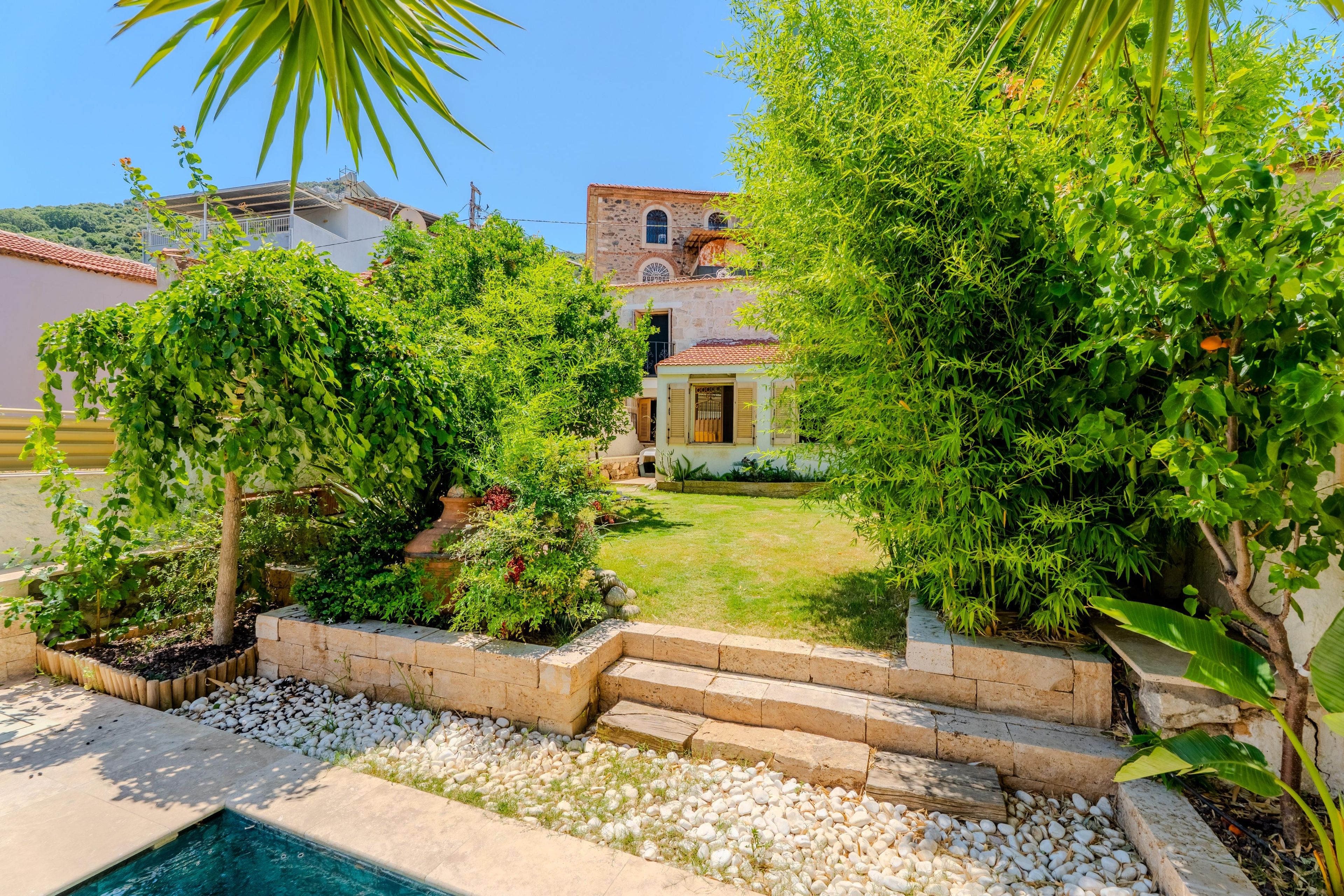 A pool surrounded by a garden with a stone pathway leading to a building with a tiled roof.