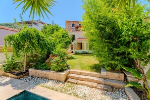 A pool surrounded by a garden with a stone pathway leading to a building with a tiled roof.