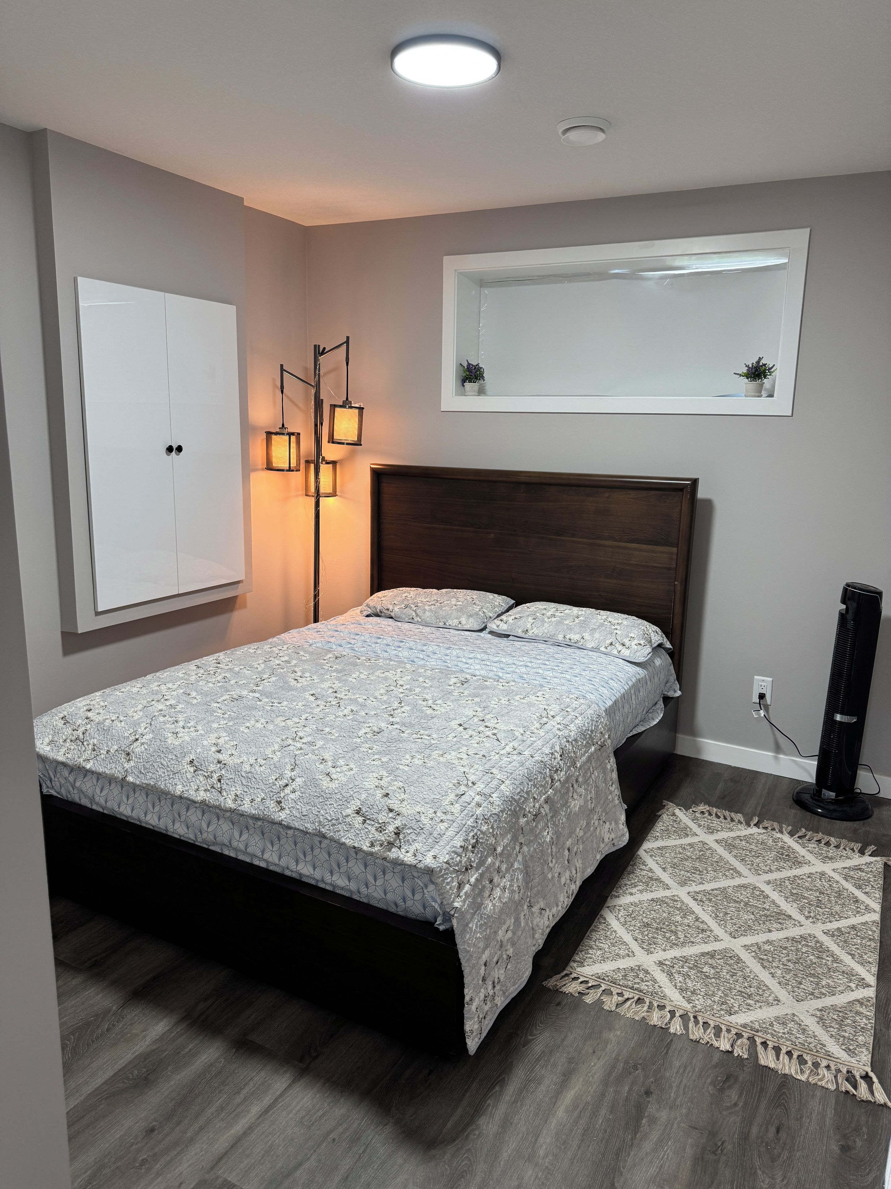 Ein Schlafzimmer mit einem Bett, einem Kleiderschrank, einer Stehlampe, einem Heizlüfter und einem Spiegel an der Wand.