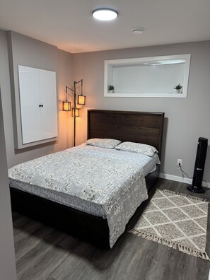 Een slaapkamer met een bed, een kledingkast, een vloerlamp, een elektrische kachel en een spiegel aan de muur.
