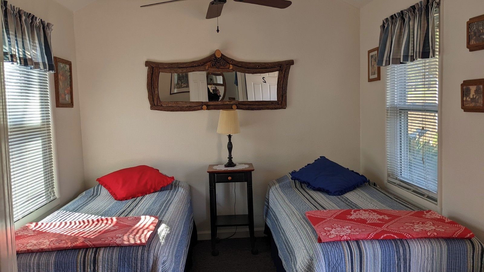 A room with two single beds, a small table with a lamp, a ceiling fan, and a mirror on the wall.