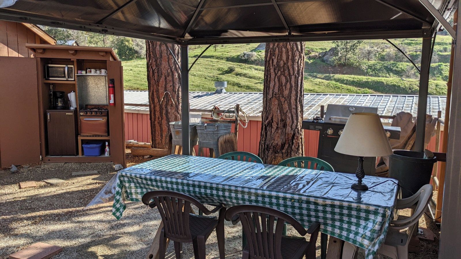 A covered outdoor dining area with a table set for two, surrounded by chairs, a lamp, and a barbecue grill.