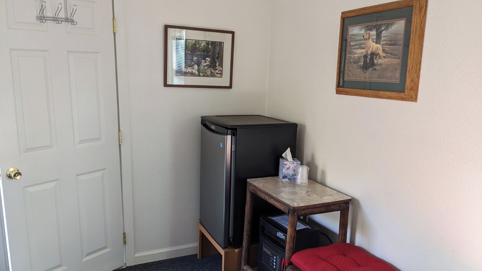 A compact room with a refrigerator, a small table with a tissue box, and two framed pictures on the wall.