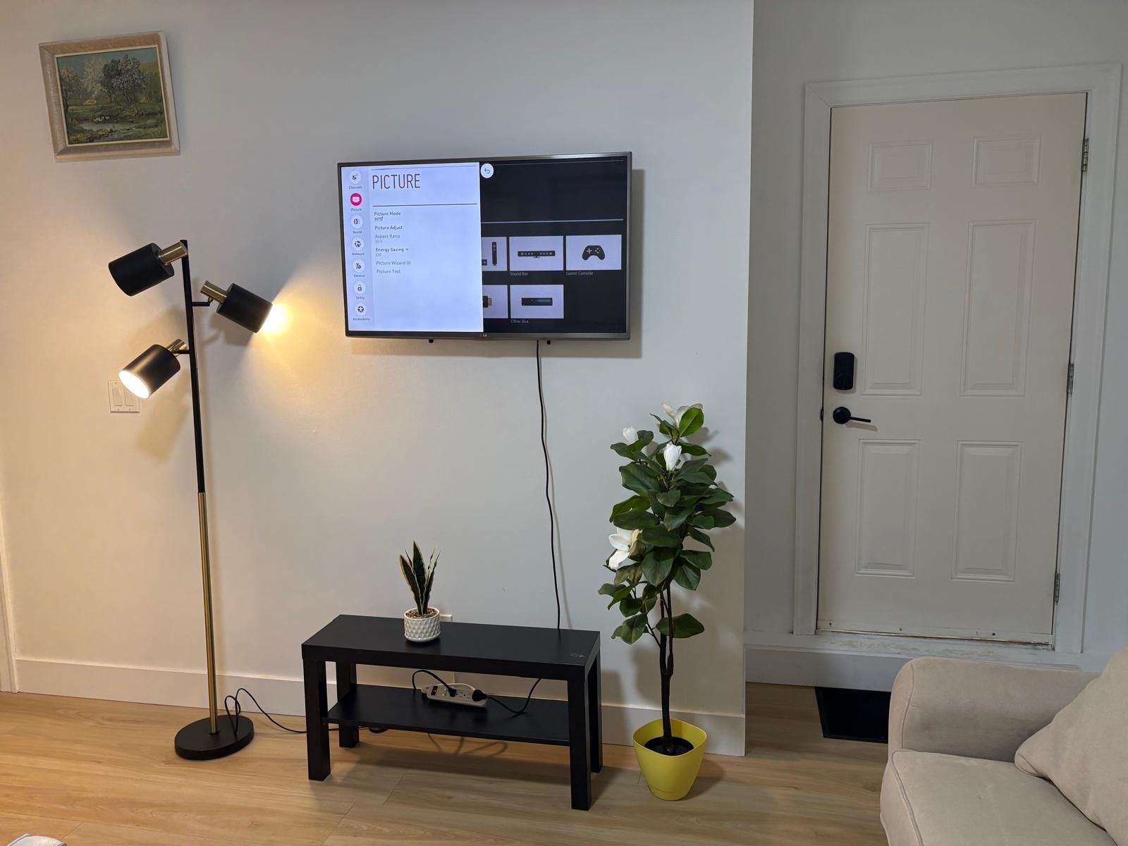 Ein Wohnzimmer mit einem an der Wand befestigten Flachbildfernseher, einer Stehlampe, einem kleinen schwarzen Tisch mit einer Pflanze und einer Tür im Hintergrund.